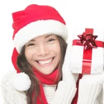 women in festive winter hat, sweater, and gloves holding a wrapped present and smiling
