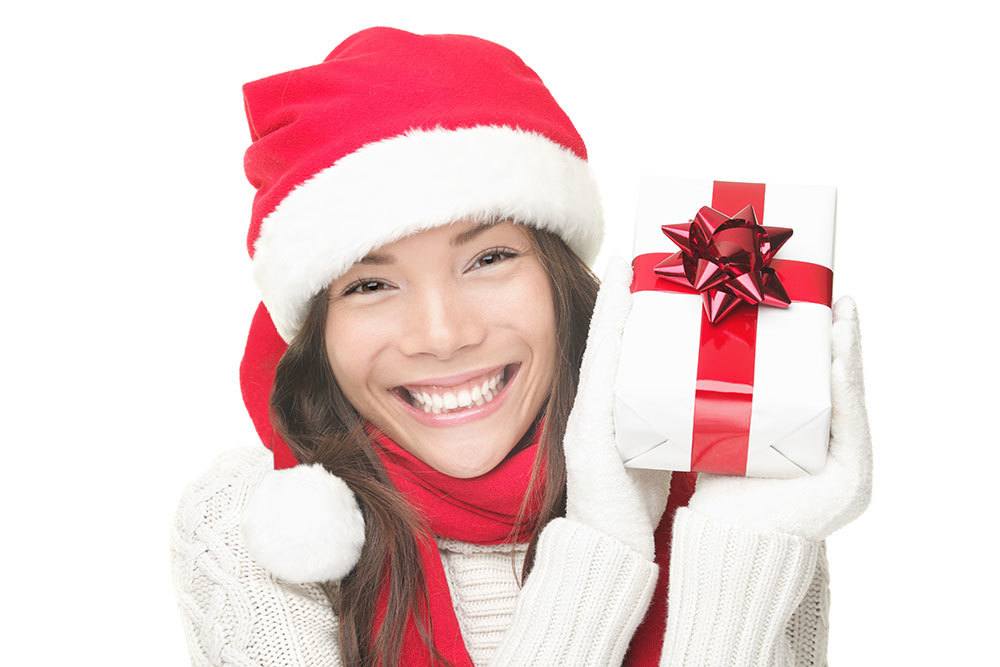 women in festive winter hat, sweater, and gloves holding a wrapped present and smiling 