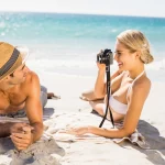 Young couple laying on a sunny beach taking photograph with the ocean in background.