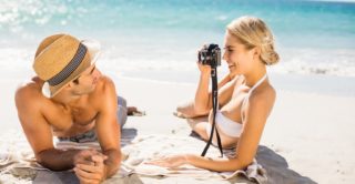 Young couple laying on a sunny beach taking photograph with the ocean in background.