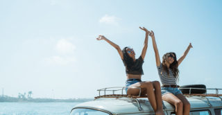 two woman waving their arms while sitting on top of VW van at the beach