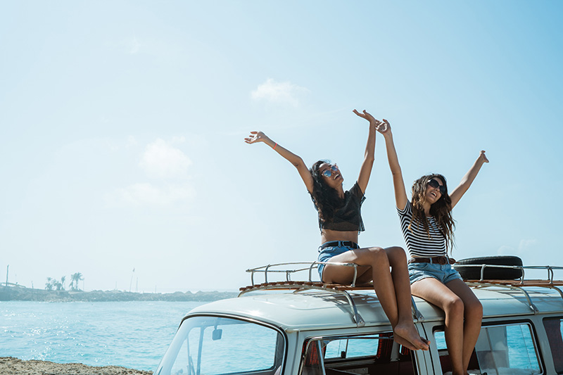 two woman waving their arms while sitting on top of VW van at the beach