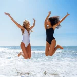 two young women smiling and jumping in the air at the beach