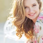 A beautiful women with long blonde hair and youthful, glowing skin, sitting outside on a sunny day wearing a floral dress