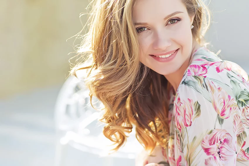 A beautiful women with long blonde hair and youthful, glowing skin, sitting outside on a sunny day wearing a floral dress