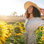 A young, beautiful women wearing a sundress and wide brim hat smiling happily in a field of sunflowers on a bright and sunny spring day