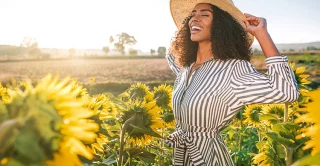 A young, beautiful women wearing a sundress and wide brim hat smiling happily in a field of sunflowers on a bright and sunny spring day
