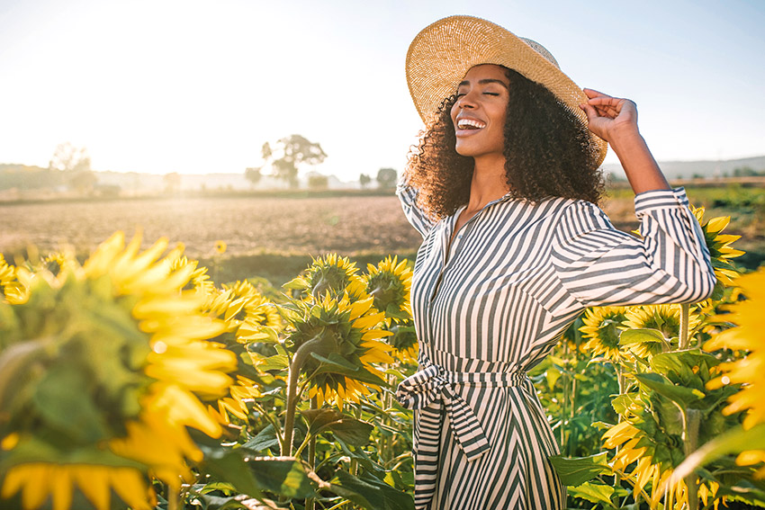 A young, beautiful women wearing a sundress and wide brim hat smiling happily in a field of sunflowers on a bright and sunny spring day