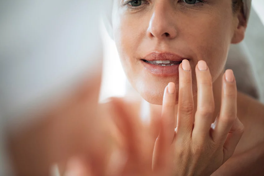 Middle-aged caucasian women looking closely into a mirror at her lips which look youthfully plump, hydrated, and wrinkle free