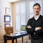 A smiling Dr. Victor Lacombe, wearing a black sweater, standing in his consultation room.