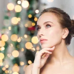 Bare-shouldered woman's head and shoulders, with hand under chin, Christmas tree background