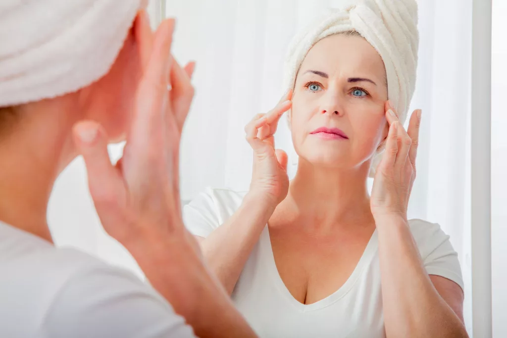 mature woman examining fine lines and wrinkles around her eyes in the mirror