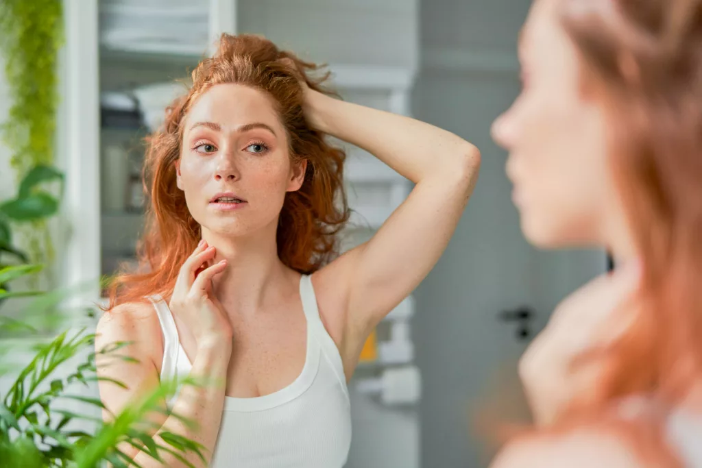 redheaded woman looking at the freckles on her face in the mirror