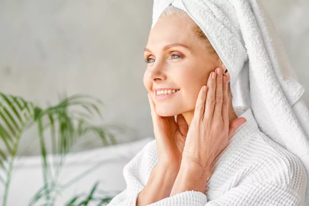 mature woman in robe and towel over hair touching her youthful-looking face