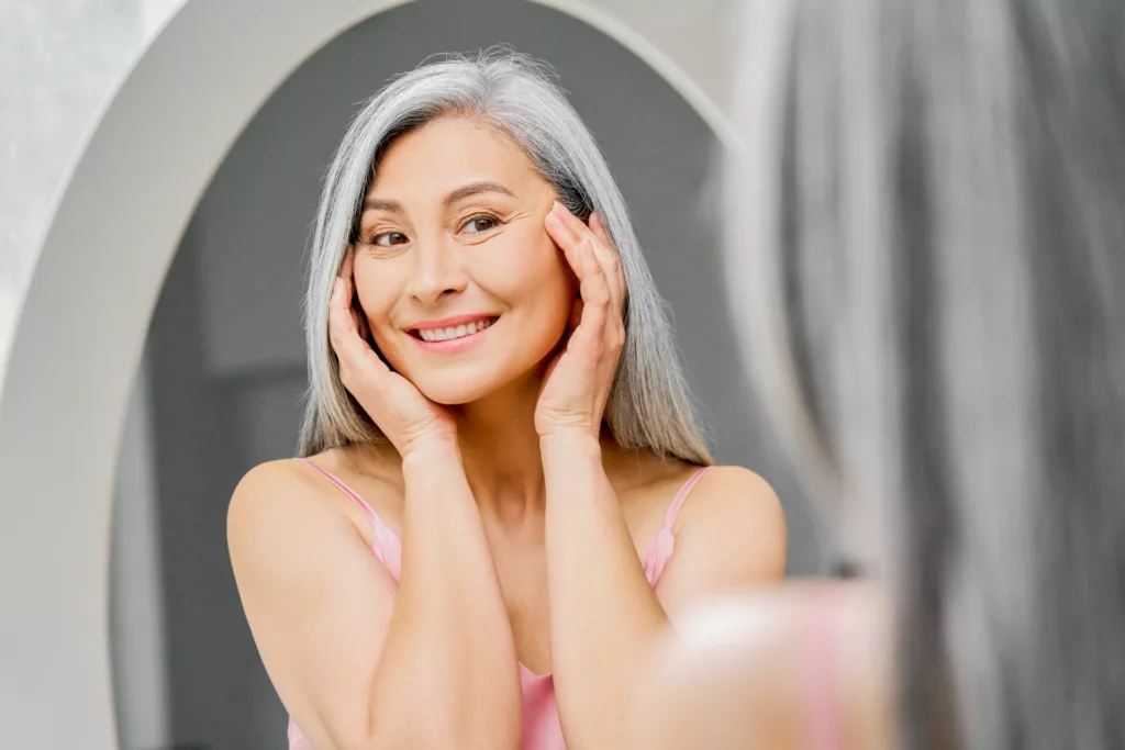 attractive mature woman admiring her youthful appearance in the mirror while cupping her hands to the sides of her face