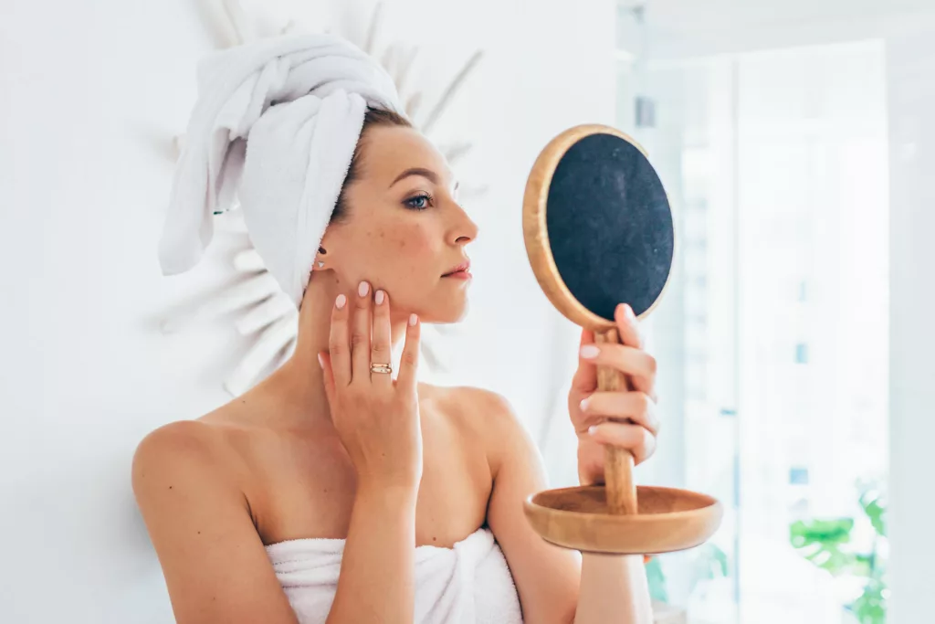 attractive woman in a bath towel with her hair up admires her strong jawline and impressive cheek contours in the mirror