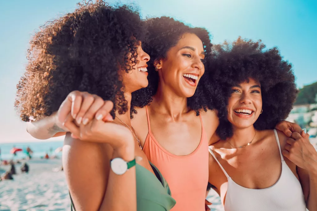 three beautiful black women with natural hair smiling and laughing at the beach while embracing one another