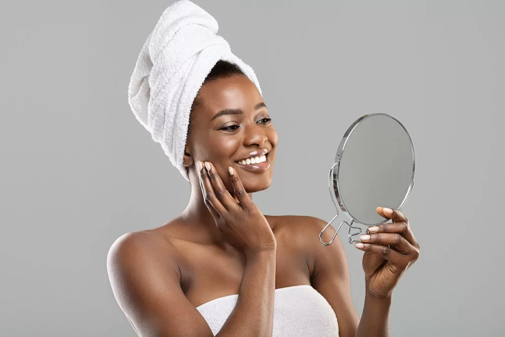 African American woman wearing towel on body and around hair, admiring her face in a hand mirror.