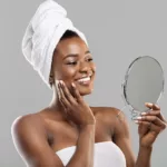African American woman wearing towel on body and around hair, admiring her face in a hand mirror.