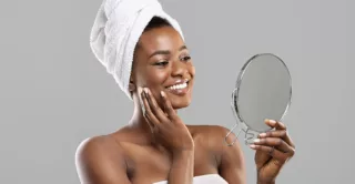 African American woman wearing towel on body and around hair, admiring her face in a hand mirror.