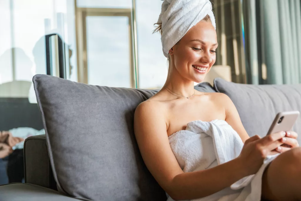young woman with gorgeous 3/4 profile sits in a towel on the sofa looking at her phone