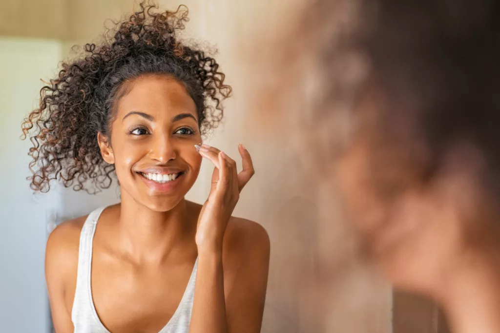 beautiful black woman with full cheeks applies moisturizer while looking at herself in the mirror