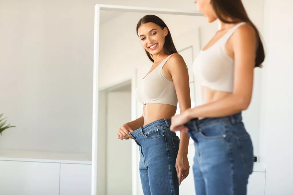 Young woman with long brown hair smiling while in front of mirror holding out waistband of too-big jeans