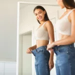 Young woman with long brown hair smiling while in front of mirror holding out waistband of too-big jeans