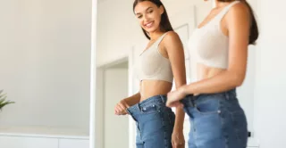 Young woman with long brown hair smiling while in front of mirror holding out waistband of too-big jeans