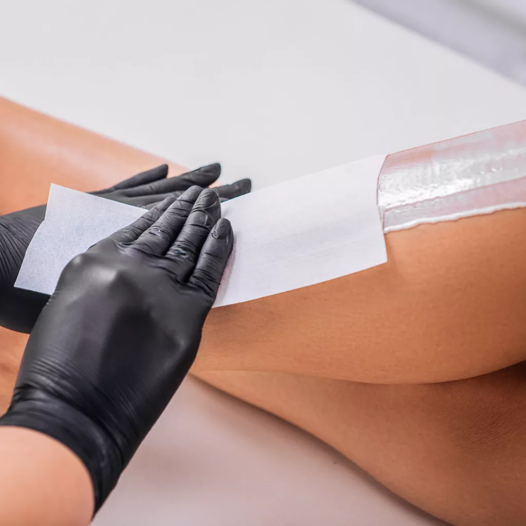 esthetician wearing gloves performs traditional waxing on patient's leg