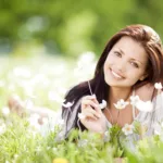 smiling woman with long brown hair lying on her stomach in a field, propped on elbows, holding a wild flower