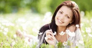 smiling woman with long brown hair lying on her stomach in a field, propped on elbows, holding a wild flower