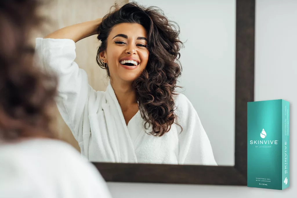 Woman pulling her hair away from her face and smiling at her mirror reflection. Box of Skinvive on shelf.