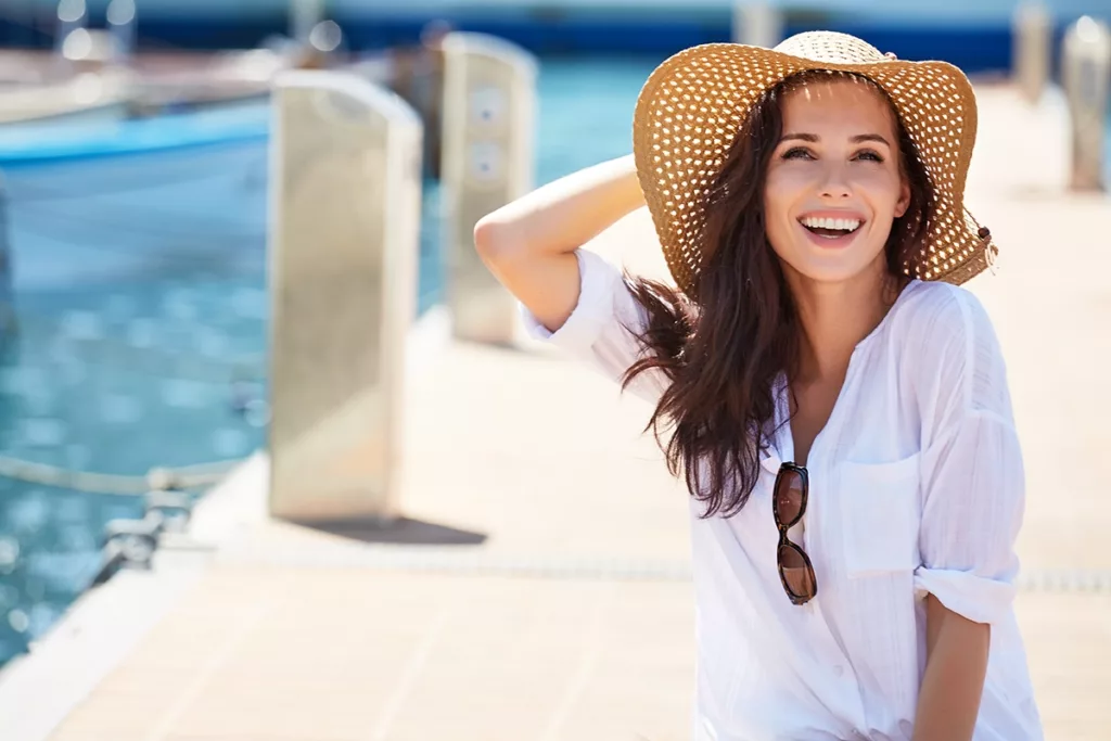 A beautiful young women with long brown hair wearing a sun hat and white linen top, sitting on a boat dock, in the sun, smiling and laughing while enjoying the view of the harbor