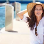 A beautiful young women with long brown hair wearing a sun hat and white linen top, sitting on a boat dock, in the sun, smiling and laughing while enjoying the view of the harbor