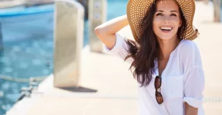 A beautiful young women with long brown hair wearing a sun hat and white linen top, sitting on a boat dock, in the sun, smiling and laughing while enjoying the view of the harbor