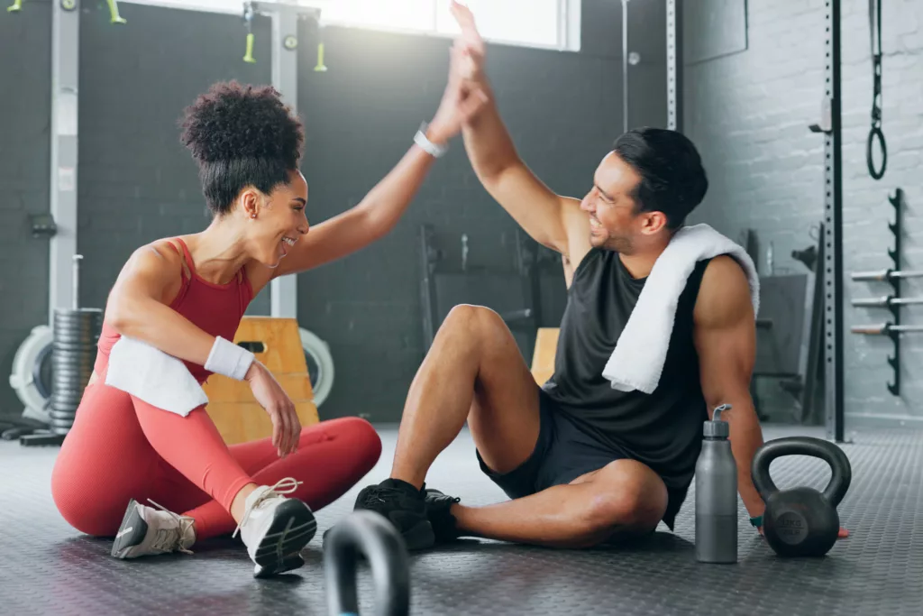 Two people sitting down in a gym after an exercise, high-fiving and laughing together; demonstrating the feeling of achievement with an Emsculpt Medical Gym membership at Artemedica