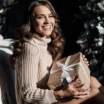A beautiful woman sitting next to an elegant Christmas tree holiday a present and smiling