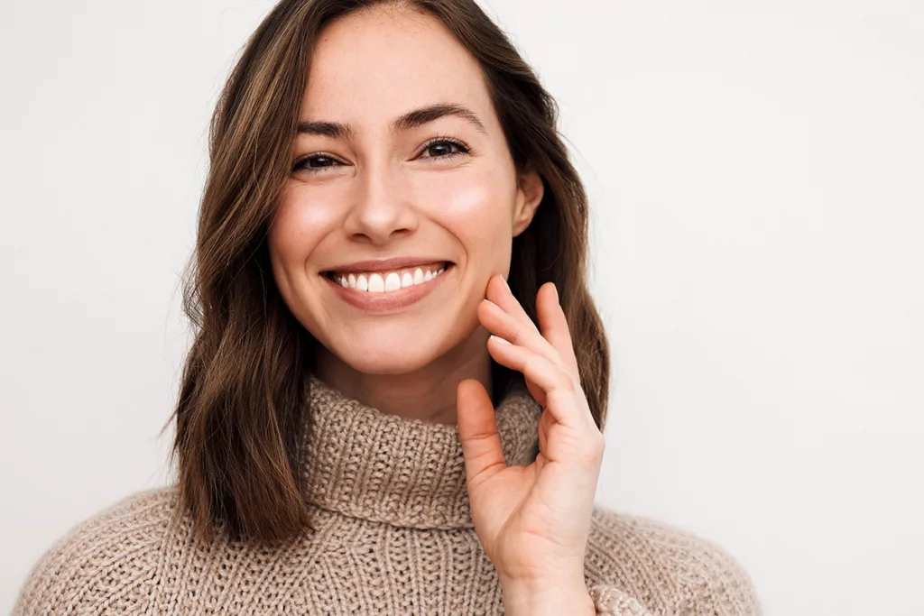 A smiling woman in a brown sweater with glowing skin