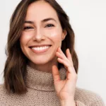 A smiling woman in a brown sweater with glowing skin