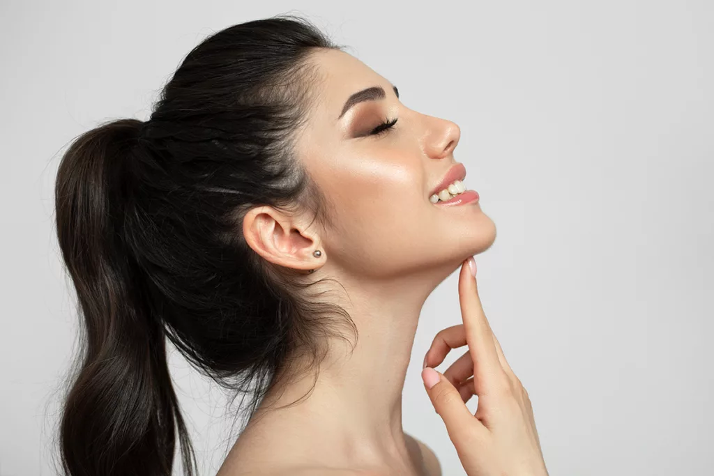 A smiling young woman in a white room, touching her chin and enjoying a well-defined jawline after EmFace Submentum non-surgical double chin treatment.