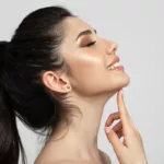 A smiling young woman in a white room, touching her chin and enjoying a well-defined jawline after EmFace Submentum non-surgical double chin treatment.