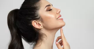 A smiling young woman in a white room, touching her chin and enjoying a well-defined jawline after EmFace Submentum non-surgical double chin treatment.