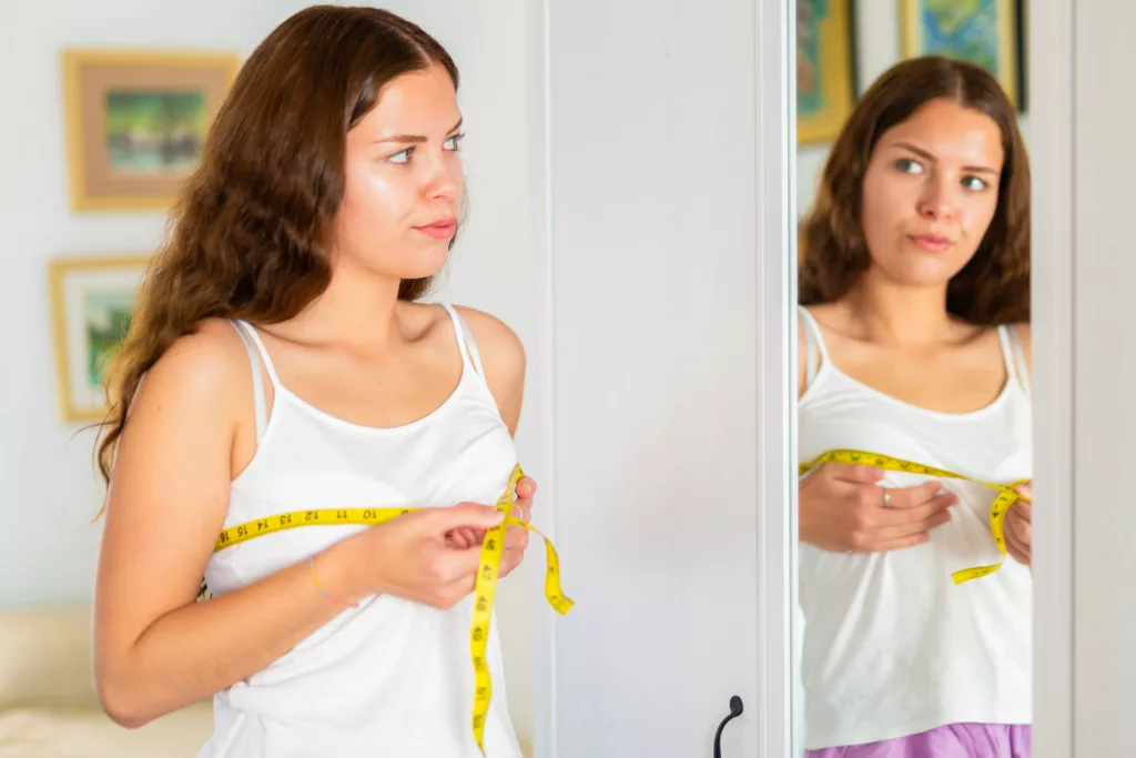 young woman measures her bustline in the mirror while contemplating breast augmentation surgery