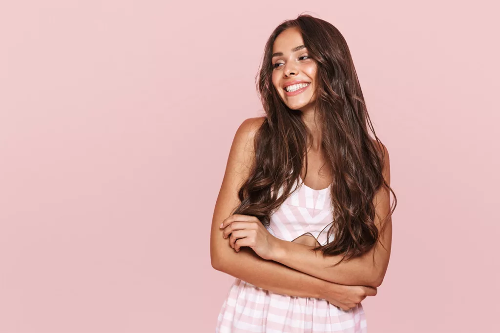 A smiling woman in a pink and white striped outfit folds her arms and stands against a pink background.