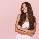 A smiling woman in a pink and white striped outfit folds her arms and stands against a pink background.