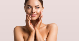 A smiling woman with clear skin holds her hands to her face, against a pink background.
