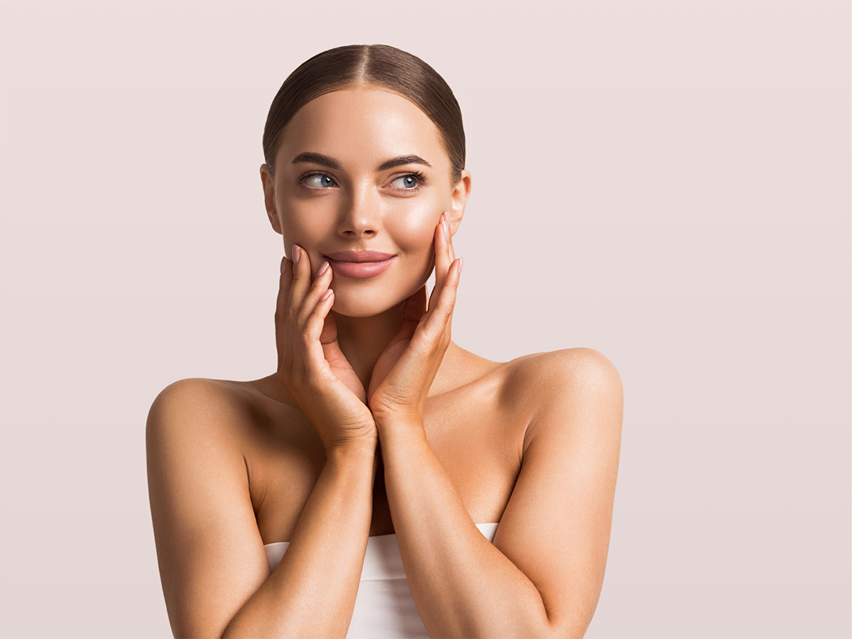 A smiling woman with clear skin holds her hands to her face, against a pink background.