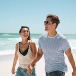 A happy young couple holds hands and walks along the beach in summer, smiling.