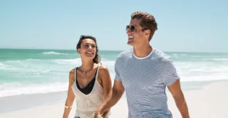 A happy young couple holds hands and walks along the beach in summer, smiling.